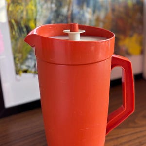 Può includere: Brocca vintage in plastica arancione con coperchio e manico bianchi. La brocca ha una superficie strutturata e un beccuccio per versare facilmente. Il manico è rettangolare e fissato lateralmente. Probabilmente utilizzato per servire bevande. Capacità di circa 2 litri.