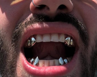 May include: A set of silver-coloured metal tooth caps designed as fangs, worn on the upper and lower canine teeth. The metallic covers have a smooth, reflective finish and are fitted over the natural teeth, creating a pointed, sharp appearance.