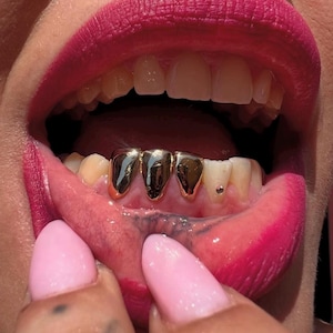 Può includere: Primo piano di una bocca con rossetto rosa acceso e denti d'oro. I denti d'oro sono un set di tre denti collegati. Le unghie sono dipinte di rosa. È visibile un piercing al labbro.