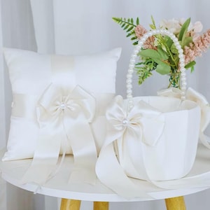 May include: Ivory satin ring bearer pillow and flower girl basket set. The pillow features a large bow with a pearl accent. The basket has a pearl handle and a matching bow. A small bouquet of flowers sits in the basket.