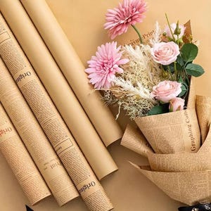 May include: A bouquet of pink flowers wrapped in newspaper-style paper, with rolls of brown paper and additional newspaper-style paper. The flowers include pink gerbera daisies and roses. The paper has printed text.