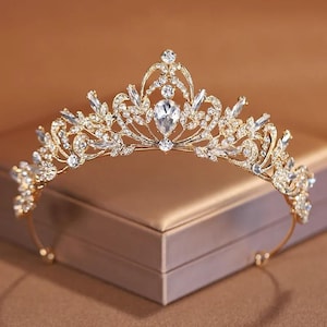 Peut inclure: Un diadème doré orné de cristaux clairs et étincelants. Le diadème présente un cristal central en forme de goutte d'eau et des motifs floraux et de feuilles complexes. La bande incurvée est ornée de petits cristaux.