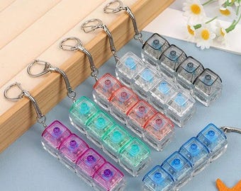 Puede incluir: Una colección de llaveros de plástico transparente y colorido. Cada llavero presenta una serie de pequeños compartimentos cuadrados con tapas de diferentes colores, incluyendo rosa, verde, rojo, azul y gris. Los llaveros están unidos a anillos metálicos plateados.