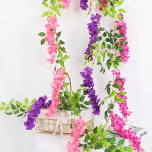 Puede incluir: Ramas de glicinia artificiales con racimos colgantes de flores rosas, moradas y magenta junto a hojas verdes. Los tallos florales caen sobre una cesta rectangular de alambre dorado con un forro de tela blanca decorativa.