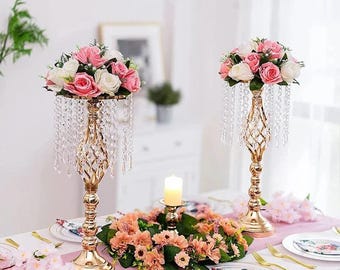 May include: Two gold-colored, ornate candle holders with hanging crystal accents, each topped with a bouquet of pink and white roses. A floral centerpiece and place settings complete the elegant table setting.