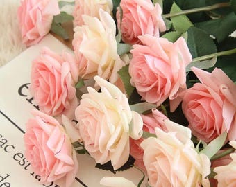 May include: A collection of artificial roses in various shades of pink and cream. The roses have green stems and leaves, and are arranged on a white surface. The image includes the text "Gekiwt".