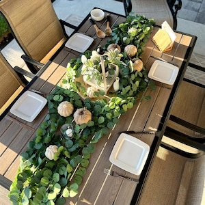 Puede incluir: Una mesa de comedor de madera para exteriores, preparada para una reunión de otoño. La mesa está decorada con una guirnalda verde, pequeñas calabazas, platos blancos y una linterna decorativa. Las sillas tienen respaldo y asiento de malla marrón.