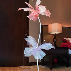 Peut inclure: Un lampadaire décoratif avec une tige blanche et deux grands abat-jours en forme de papillon. L'abat-jour supérieur est rose, et l'abat-jour inférieur est violet clair avec des accents irisés. La lampe est dans une pièce avec une porte marron foncé et une table de chevet.