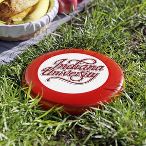Pode incluir: Um frisbee vermelho e branco com a inscrição "Indiana University" em fonte cursiva. O frisbee está sobre a relva verde. Perfeito para jogos ao ar livre.