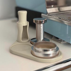 Könnte beinhalten: Ein hellbeige Kaffee-Station-Organizer mit einem Kaffeestampfer, einem Siebträgerhalter und einem Metallstampfer. Auf dem Tamper steht "La Marzocco". Die Artikel befinden sich auf einer weißen Oberfläche.