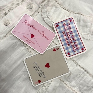 May include: Three decorative cards with text and heart designs. One pink card reads "Kindness Lingers" and "leave it everywhere you go." Another card has a blue and white gingham pattern and reads "WHAT YOU LOVE WILL ALWAYS FIND YOU BACK." The third card is gray and reads "Your heart already knows what to do. Listen."