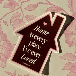 May include: A sticker in the shape of a house with the text "Home is every place I've ever Loved." The sticker is dark brown with a white border and a small pink heart. The background is a floral pattern.
