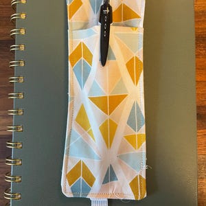 Puede incluir: Un portalápices de tela con un estampado geométrico en amarillo, azul claro y blanco, sujeto a un cuaderno verde con espiral. Un bolígrafo negro está en el soporte. El cuaderno tiene encuadernación en espiral dorada.