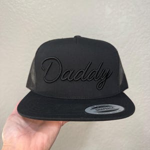 May include: A black snapback trucker hat featuring a mesh back and a solid front panel with the word Daddy embroidered in a matching black cursive script. A circular sticker is affixed to the flat brim, reading The Authentic Snapback Yupoong.