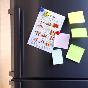 May include: A 2026 calendar with a colorful monthly layout is affixed to a black refrigerator door with blue and red magnets. Several sticky notes in yellow, pink, and green are also attached, creating a functional and organized display.