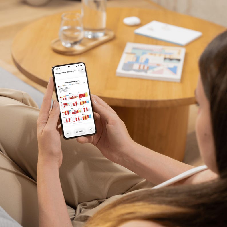 May include: A person holding a black smartphone displaying a calendar with red, white, and blue bar graphs. The phone is in focus, with a wooden table and a glass of water in the background. A book and a white wireless earbud are also visible.