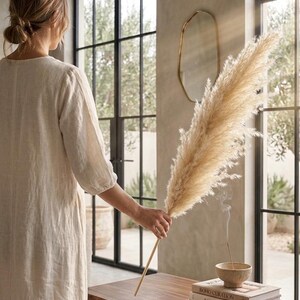 May include: A person holding a large, dried pampas grass plume. The beige plume is held over a wooden table with books and a small bowl. The room has large windows and a gold-framed mirror.