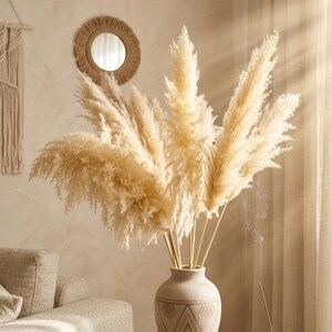 May include: A beige vase with a geometric pattern holds a large arrangement of dried pampas grass. The vase sits on a wooden table with books and an incense burner. A round mirror hangs on the wall in the background.