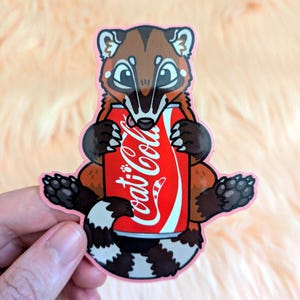 May include: A die-cut vinyl sticker featuring a brown and tan coati with a striped tail holding a red soda can with the white text Coca-Cola printed on the front. The illustration has a light pink border and dark brown paws with visible claws.