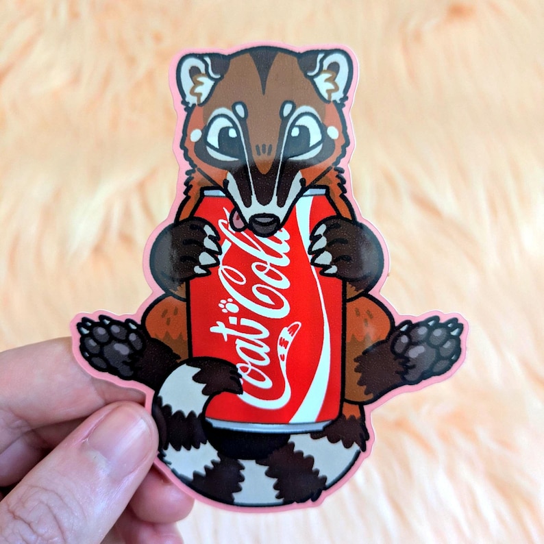 May include: A die-cut vinyl sticker featuring a brown and tan coati with a striped tail holding a red soda can with the white text Coca-Cola printed on the front. The illustration has a light pink border and dark brown paws with visible claws.