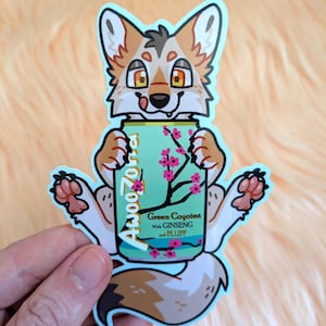May include: A die-cut sticker featuring a cartoon fox with tan and white fur holding a light green beverage can. The can displays the text AwooZona, Green CoyoTea with GINSENG and FLUFF, and a graphic of a cherry blossom branch with pink flowers.