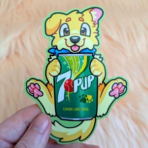 May include: A die-cut sticker featuring a yellow cartoon dog holding a green soda can. The can displays the text 7 PUP, a red ball graphic, and the words LEMON LIME SODA. The dog has pink paw pads and a blue collar, with a light green border outline.