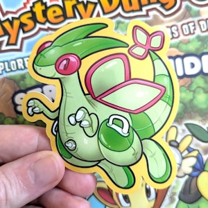 May include: A green and pink cartoon sticker of a dragon-like creature with large eyes, wings, and a tail. The sticker is on a yellow background and is being held by a hand. The sticker is from the "Mystery Dungeon" series.
