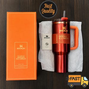 May include: A metallic red Stanley Quencher H2.0 tumbler with an orange handle and straw, accompanied by its orange retail box and a small product information card. The tumbler features a branded band with the text The Flowstate Quencher H2.0.
