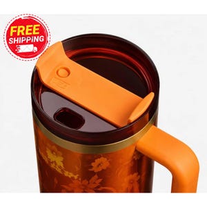 May include: A dark orange travel mug featuring a floral pattern in lighter orange and yellow tones. The mug has a dark translucent lid with a bright orange sliding closure mechanism and a matching orange handle attached to the side.