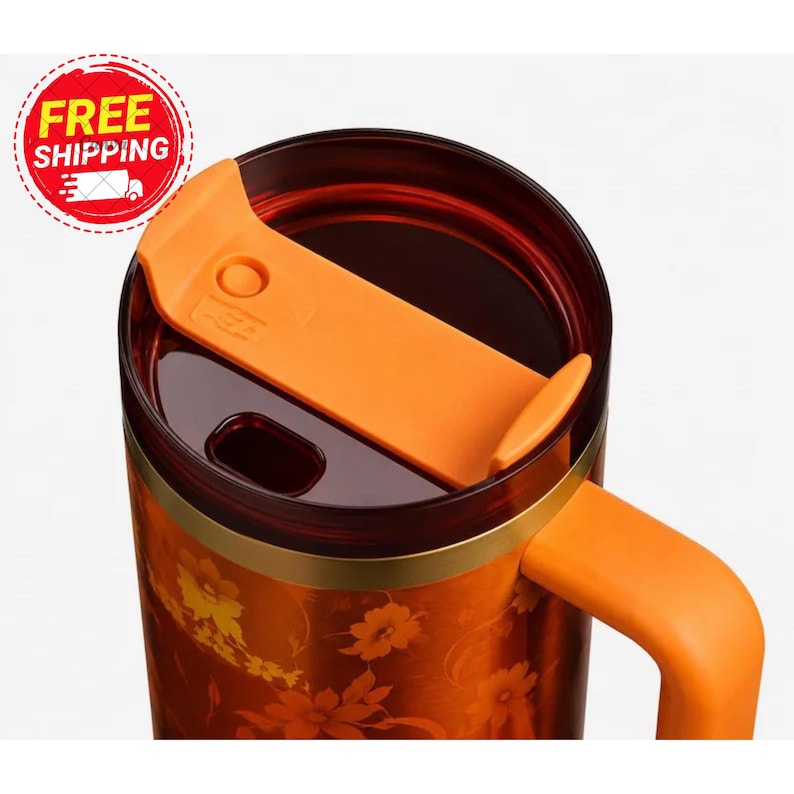 May include: A dark orange travel mug featuring a floral pattern in lighter orange and yellow tones. The mug has a dark translucent lid with a bright orange sliding closure mechanism and a matching orange handle attached to the side.
