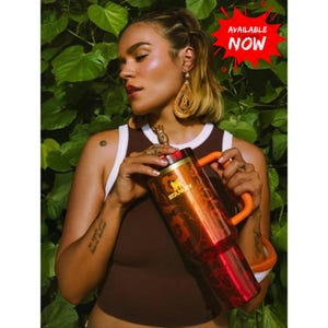 May include: A tall, orange-red Stanley tumbler with a dark floral pattern, a bright orange handle, and a gold-toned pineapple-shaped lid topper. The tumbler features the Stanley logo in gold text and a red starburst graphic with the text AVAILABLE NOW.