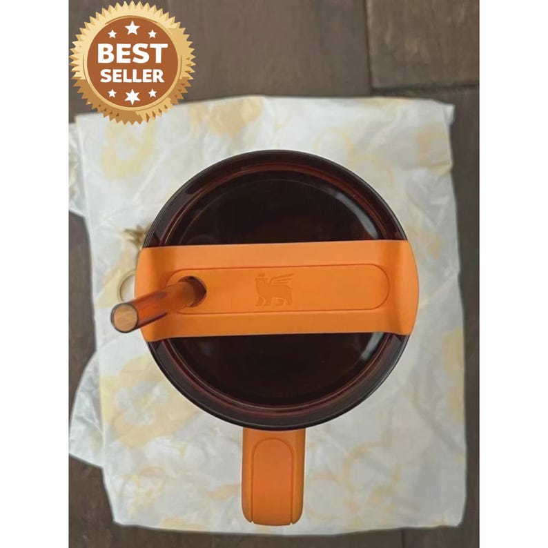 May include: A top-down view of a dark brown tumbler featuring an orange lid with a matching orange handle and a translucent brown straw. The lid displays a small embossed winged creature logo. A gold circular badge reads BEST SELLER.