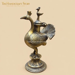 May include: A brass bird-shaped oil lamp or container featuring intricate etched feather patterns on the wings and body. The piece stands on a circular pedestal base and includes a small bird-shaped finial on the lid, with the text THE HANDICRAFT STORY.