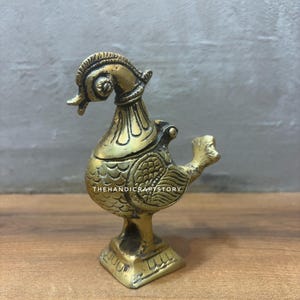 May include: A brass bird figurine with intricate detailing. The bird has a decorative headpiece and a detailed wing design. The base is square and also features detailed patterns. The text "THEHANDICRAFTSTORY" is visible on the body of the bird.