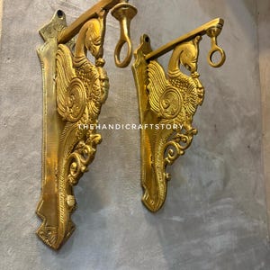 Peut inclure: Deux supports muraux en métal doré présentant un motif de paon orné avec des détails de plumes complexes et une boucle de suspension circulaire au sommet. Le texte THEHANDICRAFTSTORY est visible sur la surface des supports, qui ont une finition polie et une plaque de montage verticale.