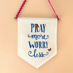 Puede incluir: Un cartel blanco con las palabras "PRAY more. WORRY less." en escritura azul, adornado con acentos de mariposa. El cartel está suspendido por una cinta rosa y un palillo de madera, creando una decoración de pared.