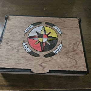 May include: Wooden box with a Native American design. The lid features a circular emblem with a sun-like symbol in red, yellow, black, and white, surrounded by four feathers. The box has a natural wood grain finish and black trim.