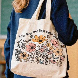 May include: A cream-colored tote bag featuring a floral design with pink, yellow, and peach flowers, green leaves, and butterflies. The text "teach them, love them, watch them" is printed across the top. The bag has two cream-colored straps.