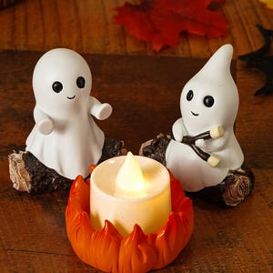 Peut inclure: Une décoration d'Halloween avec deux figurines de fantômes blancs aux yeux noirs et sourires, posées sur des socles en bois. Une bougie à piles dans un support orange en forme de flamme est devant. Des feuilles décoratives et une toile d'araignée complètent la scène.