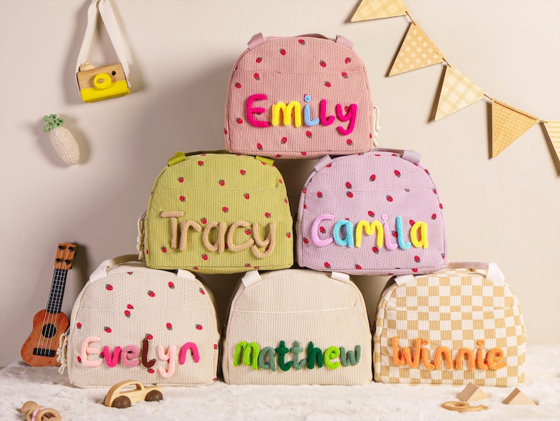 Pu&ograve; includere: Una collezione di zaini in velluto a coste in vari colori, ognuno decorato con un nome in lettere colorate in rilievo. Gli zaini presentano un motivo a fragole e sono impilati. Sono visibili anche un piccolo ukulele e un'auto giocattolo.