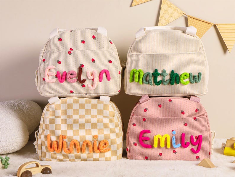Pu&ograve; includere: Quattro zaini per bambini in vari modelli e colori. Uno &egrave; beige con fragole e il nome "Evelyn" in lettere rosa. Un altro &egrave; beige con il nome "Matthew" in lettere verdi. Un terzo &egrave; a scacchi con il nome "Winnie" in arancione. L'ultimo &egrave; rosa con fragole e il nome "Emily".