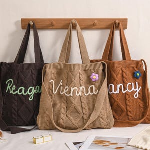 Puede incluir: Tres bolsos tote de punto trenzado en marrón, beige y marrón oscuro, cada uno con un nombre bordado en un color contrastante. Los bolsos tienen correas largas y están decorados con pequeños adornos florales. Los bolsos están colgados en un perchero de madera.