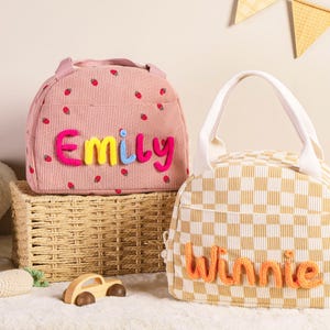 Pu&ograve; includere: Due borse per il pranzo. Una rosa con dettagli di fragole e il nome "Emily" in filato colorato, e l'altra a scacchi con il nome "Winnie" in filato arancione. Entrambe hanno manici e sono su una superficie bianca.