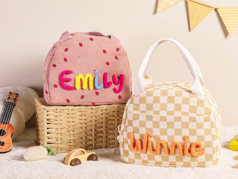 Pu&ograve; includere: Due borse per il pranzo. Una rosa con dettagli di fragole e il nome "Emily" in filato colorato, e l'altra a scacchi con il nome "Winnie" in filato arancione. Entrambe hanno manici e sono su una superficie bianca.