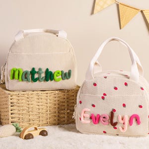 Pu&ograve; includere: Due borse per il pranzo in velluto a coste beige. Una borsa ha il nome "Matthew" in filo verde, e l'altra ha un motivo a fragole e il nome "Evelyn" in filo rosa. Entrambe le borse hanno manici bianchi.