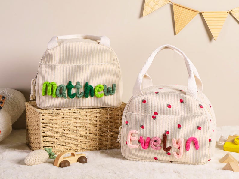 Pu&ograve; includere: Due borse per il pranzo in velluto a coste beige. Una borsa ha il nome "Matthew" in filo verde, e l'altra ha un motivo a fragole e il nome "Evelyn" in filo rosa. Entrambe le borse hanno manici bianchi.
