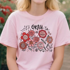 Op de afbeelding: Lichtroze T-shirt met de tekst "it's OKAY to" boven een bloemenontwerp. De bloemen zijn in rood, roze en oranje tinten, met positieve boodschappen. Het T-shirt heeft een ronde hals en korte mouwen.