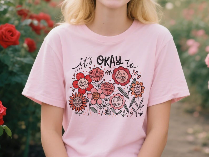 Op de afbeelding: Lichtroze T-shirt met de tekst "it's OKAY to" boven een bloemenontwerp. De bloemen zijn in rood, roze en oranje tinten, met positieve boodschappen. Het T-shirt heeft een ronde hals en korte mouwen.