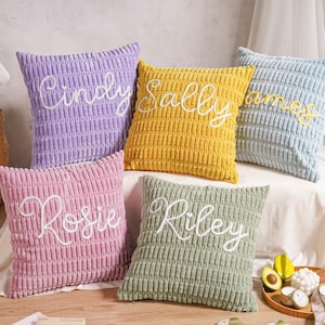 May include: Five corduroy cushions in shades of lavender, yellow, light blue, pink, and green. Each cushion features a name embroidered in white cursive: Cindy, Sally, James, Rosie, and Riley. The cushions are displayed on a white surface.