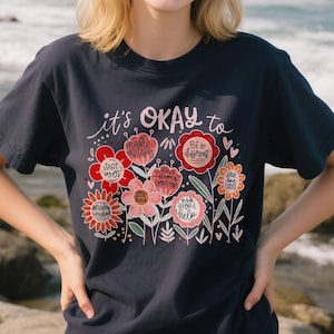 Op de afbeelding: Marineblauwe t-shirt met een bloemenontwerp en de tekst "it's OKAY to" boven de bloemen. De bloemen zijn in tinten rood, roze en oranje, met zinnen als "make mistakes" en "be different".